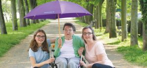Ältere Dame im Rollstuhl mit Mädchen und jüngerer Frau unter einem großen Schirm