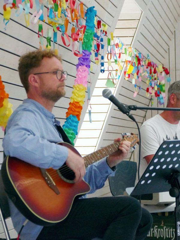Gitarrenspieler in dekoriertem Raum, im Hintergrund weiterer Musiker am Klavier